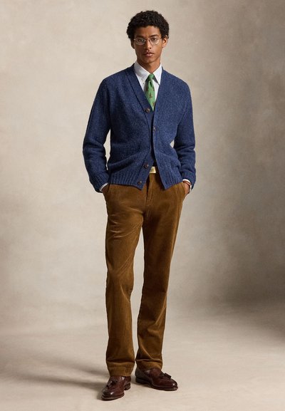 Νεαρός άνδρας με γυαλιά, μπλε ζακέτα, λευκό πουκάμισο με πράσινη γραβάτα, καφέ παντελόνι από κορδόνι και καφέ παπούτσια με φούντες, στέκεται με τα χέρια στις τσέπες.