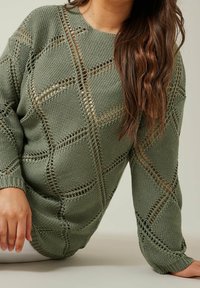 Kobieta w oliwkowym, dzianinowym swetrze z ażurowym wzorem w romby, z długimi falującymi brązowymi włosami, opierająca jeden rękę na jasnej powierzchni.