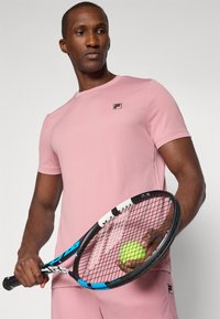 Lys rosa treningsskjorte med korte ermer, rund halslinning og F-logo. Holder en svart tenniskølle med en grønn tennisball.