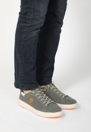 Graue Leinensneaker mit orangen Akzenten und hellen Sohlen. Mit runden Schnürsenkeln und einem Logo-Patch an der Seite. Getragen mit dunklen Jeans.