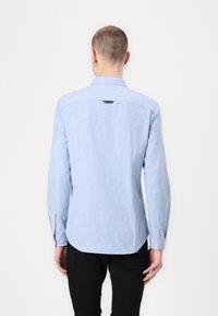 Person med kort hår som bär en ljusblå långärmad skjorta och svarta byxor, stående med ryggen mot betraktaren mot en enfärgad bakgrund.