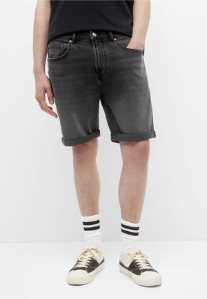 Personne portant un short en jean noir retroussé, des chaussettes blanches à rayures noires et des baskets crème et noires, debout devant un fond blanc.