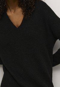 Pull en tricot noir avec un col en V, manches longues et une texture douce, mettant en valeur une silhouette simple et moderne.