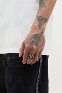 Anillo de metal negro con un diseño detallado de un pájaro, llevado en una mano con un gran tatuaje de calavera. La mano descansa sobre unos pantalones oscuros y una camisa blanca.