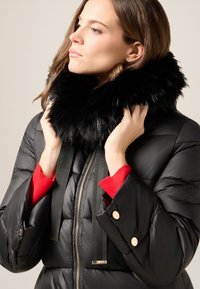 Chaqueta acolchada negra con cuello de piel sintética negro, que presenta un cierre de cremallera, tirantes de tela negra y acentos de manga de punto rojo.