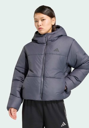 Jonge vrouw draagt een donkergrijze donsjas met capuchon en zwarte broek, staand met één hand op haar heup.