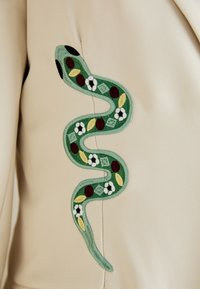 Design brodat cu un șarpe verde, accentuat cu roșu și galben, având motive florale pe un fundal de țesătură bej.