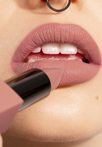 Matte, mauve leppestift påføres fyldige, veldefinerte lepper med en subtil glans. Leppestiften har en svart og myk rosa rørdesign.