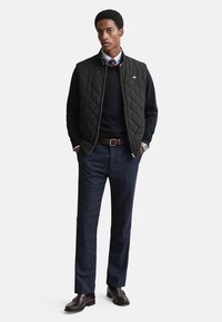 Schwarze gesteppte Weste über einem marineblauen Pullover, kombiniert mit maßgeschneiderten marineblauen Hosen. Brauner Ledergürtel und Schuhe. Hellblaues Hemd mit gemusteter Krawatte.