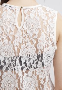 Top de encaje blanco con patrones florales, textura translúcida, cuello redondo y detalle de botón en la espalda. Diseño sin mangas.