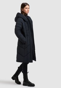 Jonge vrouw draagt een lange zwarte donsjas met capuchon, zwarte leggings en zwarte stevige laarzen, staand tegen een lichtgrijze achtergrond.