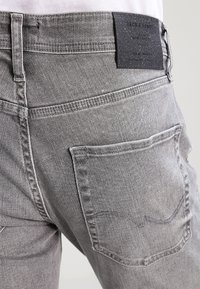 Grå denimjeans med en bakficka och en kontrasterande svart tygetikett vid midjan. Slät textur och subtila blekna detaljer.