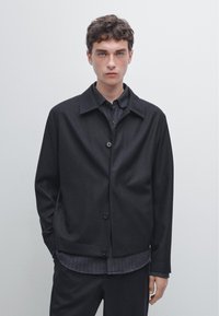 Czarna kurtka na guziki z płaskim kołnierzem, wykonana z fakturowanej tkaniny o luźnym kroju, warstwowo ułożona na ciemnej koszuli w paski.