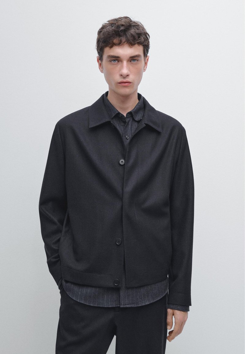 Czarna kurtka na guziki z płaskim kołnierzem, wykonana z fakturowanej tkaniny o luźnym kroju, warstwowo ułożona na ciemnej koszuli w paski.