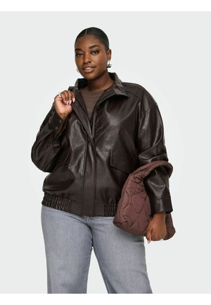 Femme portant une veste en cuir marron foncé et un jean bleu clair, tenant un sac à main marron matelassé sur un fond uni.