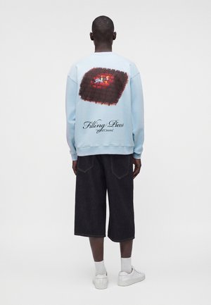 Homme portant un sweat-shirt bleu clair avec un graphisme représentant une personne sur un canapé rouge et le texte "Filling Pieces Art of Cinema" dans le dos.