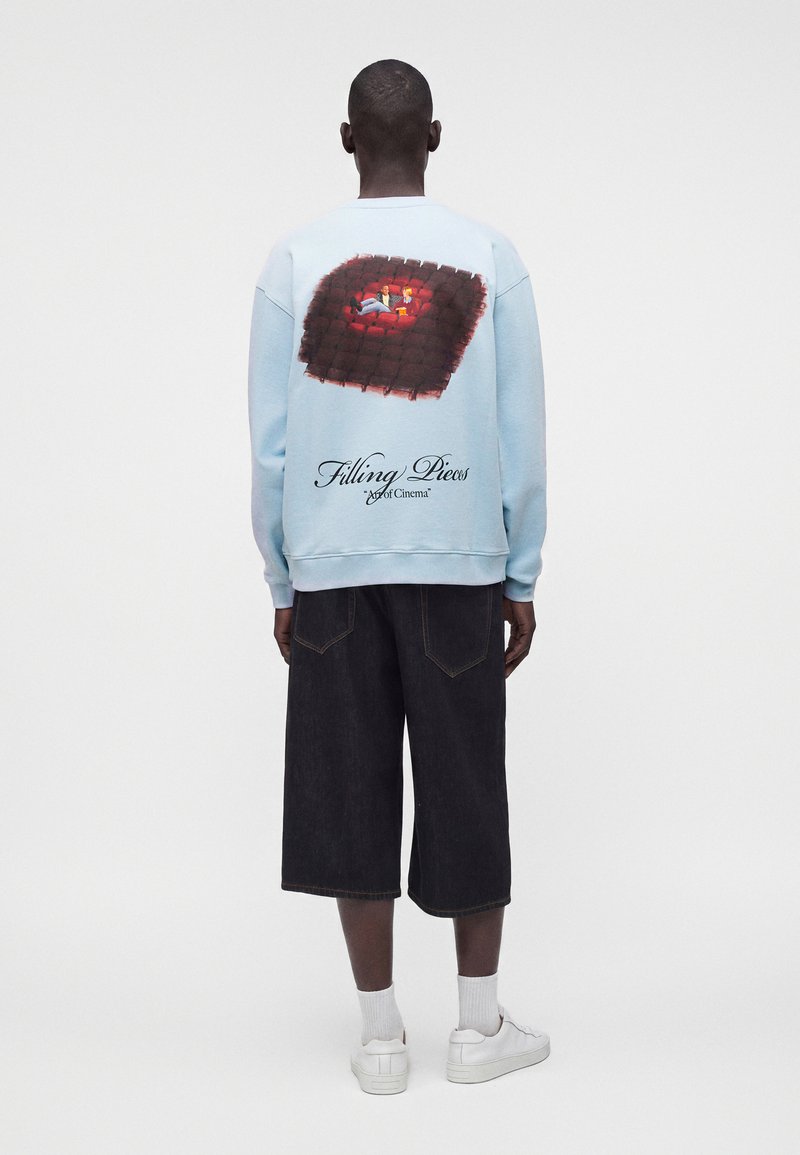 Homme portant un sweat-shirt bleu clair avec un graphisme représentant une personne sur un canapé rouge et le texte "Filling Pieces Art of Cinema" dans le dos.