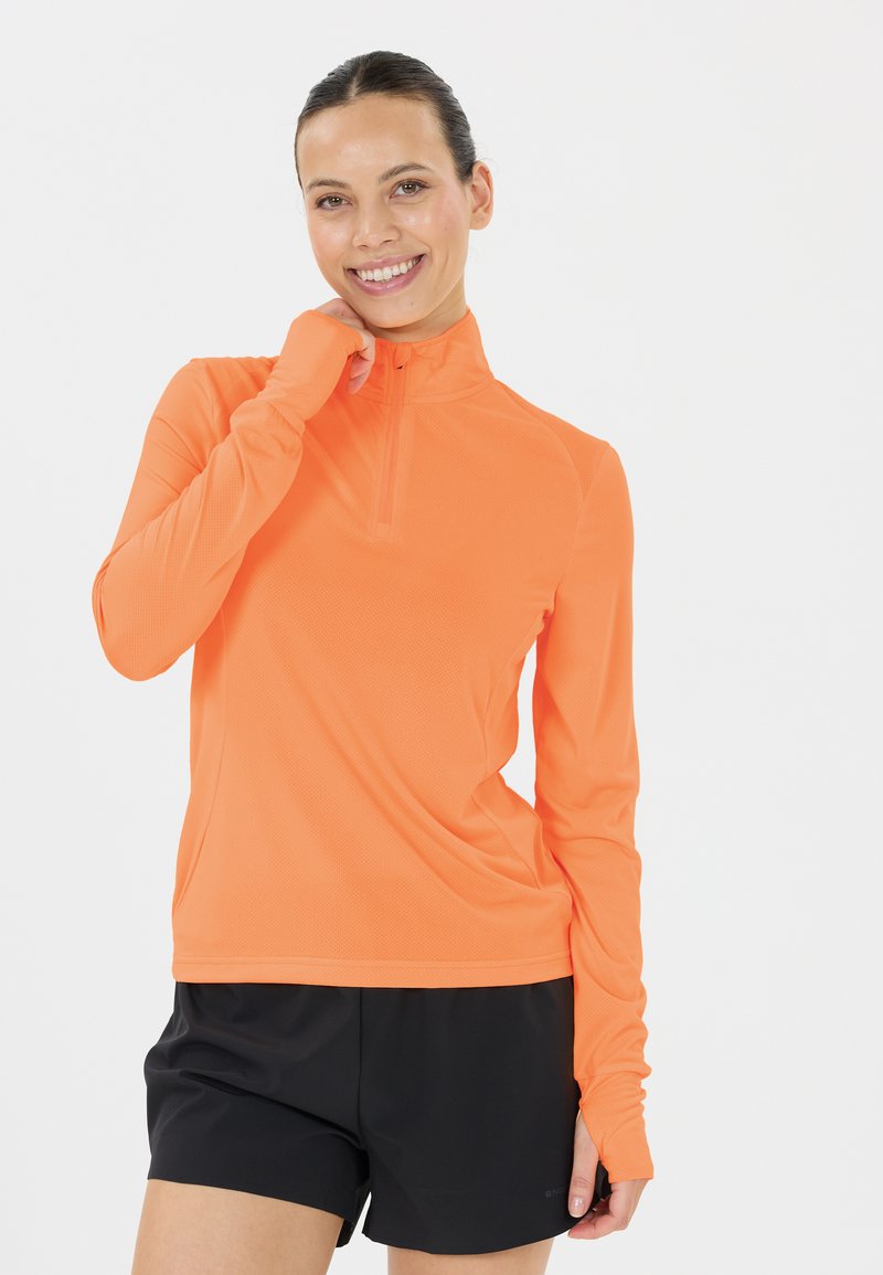 Lys orange atletisk halv-zipped trøje med lange ærmer, lavet af letvægts, tekstureret stof. Bæres med sorte shorts, der har tommelhuller.