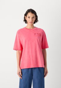 Rosa bomulls oversized t-skjorte med korte ermer, rund hals og en liten logotrykk på venstre side. Glatt tekstur, avslappet passform.