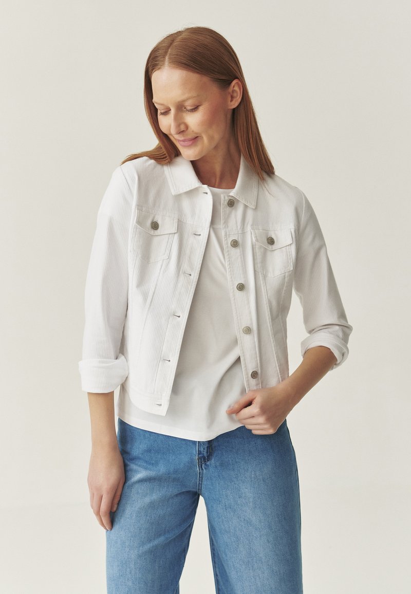 Veste en denim blanche avec un col, boutons avant visibles, deux poches poitrine et manches retroussées, portée avec un haut blanc et un jean bleu.