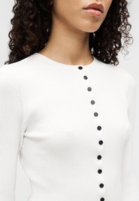 Λευκό τοπ από ριμπ πλεκτό ύφασμα με στρογγυλή λαιμόκοψη και σειρά από μαύρα κουμπιά στην μπροστινή πλευρά για αντίθεση. Μαλακή υφή, εφαρμοστή σχεδίαση.