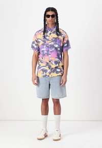 Cămașă cu mânecă scurtă și nasturi, în împrimare abstractă de violet și galben, asortată cu șorturi din denim deschis și adidași albi cu accente roșii.