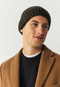 Knit beanie in dark brown with a ribbed texture. Model wears a brown coat over a black sweater with a white collar detail. Neutral background.