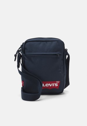 Kompaktowa granatowa torba na ramię Levi's z przednią kieszenią na zamek, regulowanym paskiem i czerwoną naszywką z logo na jednolitym jasnym tle.