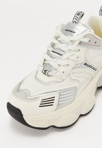 Baskets blanches et argentées avec une tige en mesh, languette rembourrée et semelle épaisse. Présente une marque, des lacets et des accents texturés.