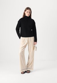 Juodas megztas turtlenecko tipo džemperis, derinamas su blizgučiais smėlio spalvos plačiais kelniais, papildomai su mažai balta rankine ir juodais sandalais.