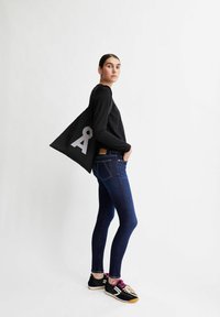 Junge Frau in dunkler Jeans, schwarzem Pullover und Turnschuhen, die eine schwarze Tragetasche mit einem großen weißen Schlüsselloch-Symbol über der Schulter trägt.