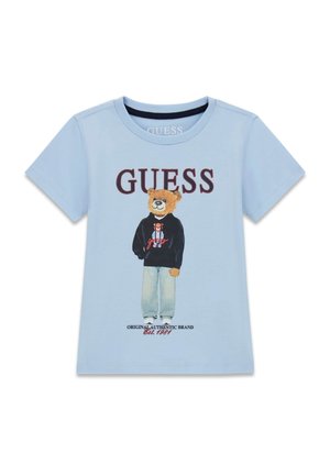 Chemise bleu clair à manches courtes représentant un ours debout portant un sweat à capuche noir, un jean et des baskets blanches avec le texte "GUESS" au-dessus et le logo en dessous.