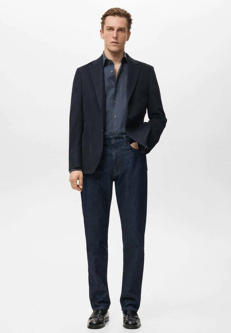 Mann steht mit einem dunklen Blazer, einem dunkelblauen, geknöpften Hemd, dunklen Jeans und schwarzen Schuhen, eine Hand in der Tasche, vor schlichtem Hintergrund.