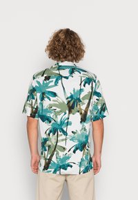 Hombre con pelo rizado que lleva una camisa de manga corta blanca con estampado de palmeras azules y verdes, pantalones beige, de pie contra un fondo liso.