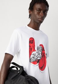 Junger Mann trägt ein weißes T-Shirt mit roten Boxhandschuhen und einer Boxer-Illustration und hängt sich eine schwarze Sporttasche über die Schulter.