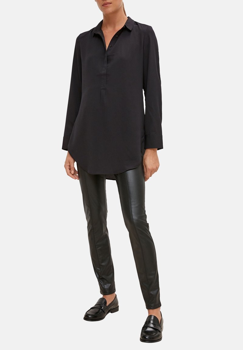 Femme portant une tunique noire à manches longues, un pantalon en cuir noir et des mocassins noirs, debout devant un fond uni.