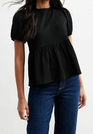 Femme portant un chemisier texturé noir à manches courtes avec un ourlet péplum ample et un jean bleu foncé sur un fond uni.