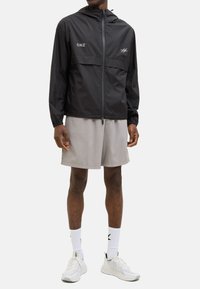 Veste légère noire avec capuche, poignets élastiques et fermeture éclair frontale. Présente un logo contrastant et une poche. Portée avec un short gris et des chaussures blanches.
