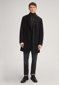 Zwarte wollen jas met inkepingen in de revers, een enkelvoudige sluiting en twee voorzakken, gedragen over een zwarte hoogsluitende binnenlaag.