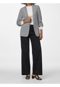 Blazer en blanco y negro con estampado de houndstooth y mangas enrolladas, superpuesto a una camisa clara, combinado con jeans de pierna amplia negros y sandalias de tacón cuadrado.