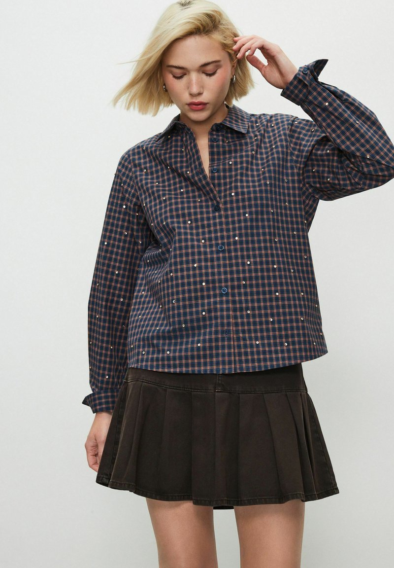 Chemise à carreaux bleue marine à manches longues avec des accents de paillettes dispersés, fermeture par boutons, associée à une jupe plissée foncée. Tissu texturé, coupe décontractée.