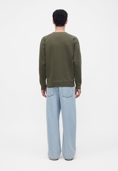 Homme aux cheveux courts et foncés portant un sweat-shirt vert olive et un jean bleu clair ample, debout de dos devant un fond uni.
