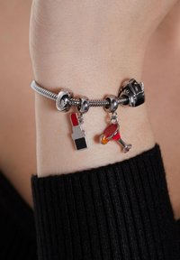 Silver charm bracelet on wrist with charms shaped like red lipstick, cocktail glass with orange garnish, and a handbag.