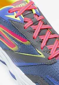 Chaussure de sport grise avec une tige en mesh texturé. Lacets et accents rouge vif, éléments de design jaune et bleu. Semelle blanche épaisse.