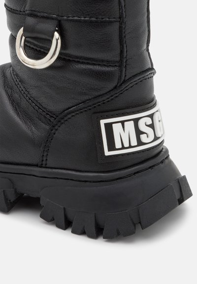 Botas de couro pretas com uma superfície texturizada, apresentando um anel de metal prateado e um patch de logó diferente. Sola grossa e resistente com padrão de aderência definido.
