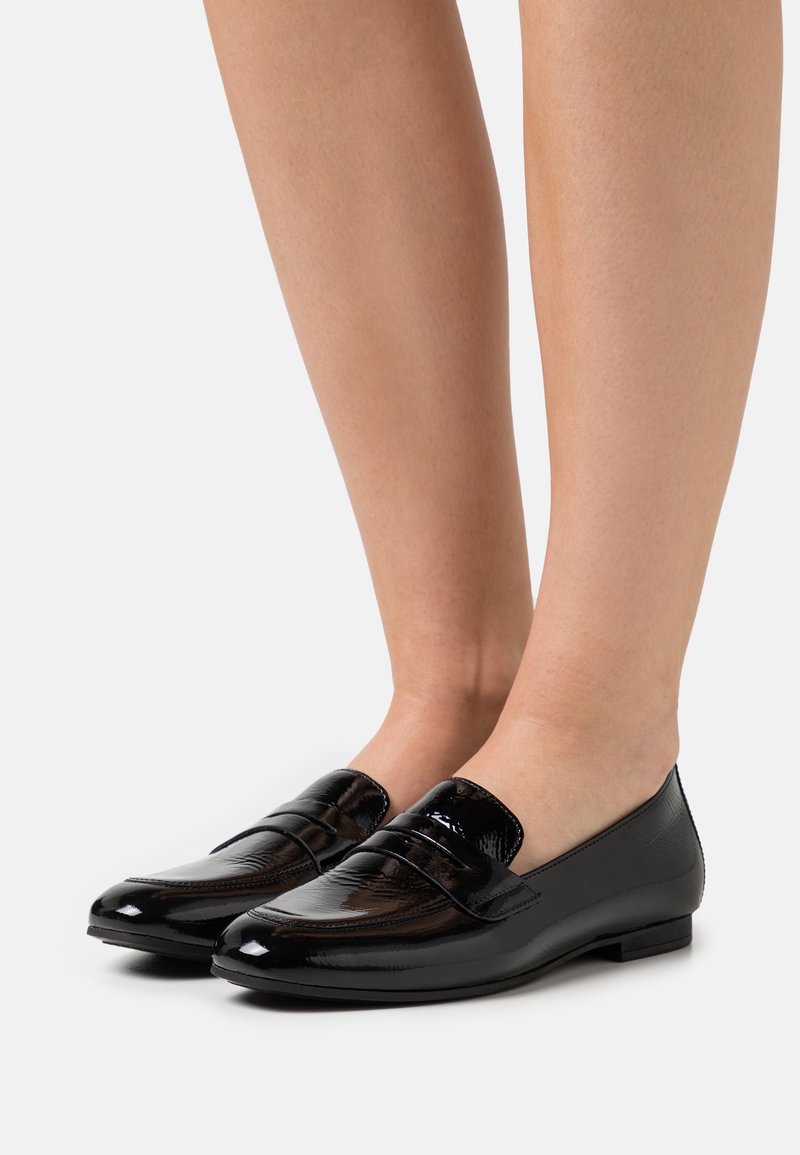Mocassins en cuir verni noir brillants portés pieds nus, présentant un talon bas élégant et un détail de sangle à l'avant.