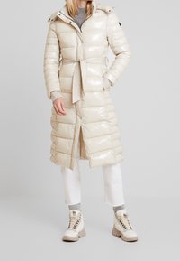 Abrigo acolchado beige largo y brillante con cintura ceñida mediante cinturón, capucha y botones a presión. Presenta un diseño acolchado y dos bolsillos. Combinado con pantalones blancos y botas.