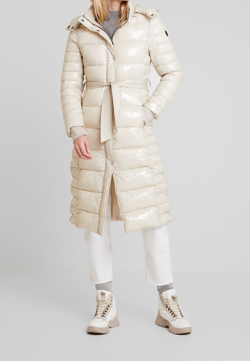 Abrigo acolchado beige largo y brillante con cintura ceñida mediante cinturón, capucha y botones a presión. Presenta un diseño acolchado y dos bolsillos. Combinado con pantalones blancos y botas.