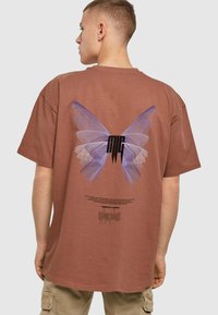 Brun oversized t-shirt med et stort lilla sommerfugl-grafik på bagsiden, med sort tekst og et subtilt tekstureret design.