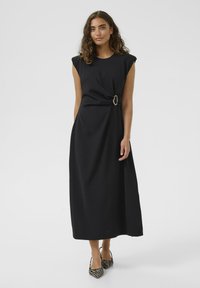 Robe maxi noire avec des manches courtes et un détail avant torsadé mettant en avant une grande boucle circulaire en argent. Tissu lisse avec une silhouette fluide.